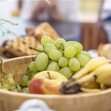 Ein Holzkorb mit frischem Obst, darunter grüne Trauben, einen Apfel und Bananen. Im Hintergrund sind weitere Lebensmittel zu sehen.