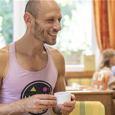 Ein lächelnder Mann hält eine Tasse in einem hellen Raum mit einem gemütlichen Ambiente. Im Hintergrund sind andere Personen am Tisch zu sehen.