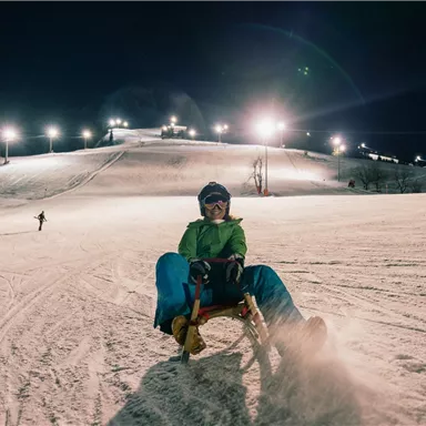 Ein Junge fährt mit einem Schlitten den beleuchteten Hang hinunter. Im Hintergrund sind weitere Wintersportler und die schneebedeckte Landschaft zu sehen.