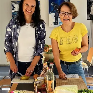 Zwei Frauen stehen an einem Tisch und bereiten frisches Gemüse vor. Der Tisch ist mit verschiedenen Lebensmitteln und Kochutensilien bedeckt.