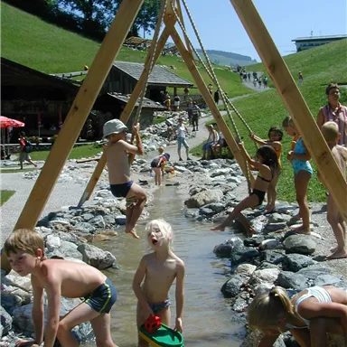 Ein Spielplatz mit Kindern, die im Wasser und auf Steinen spielen. Im Hintergrund sind Bäume und ein Gebäude sichtbar.
