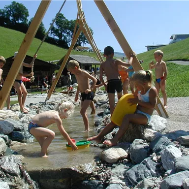 Kinder spielen in einem Wasserbereich im Freien. Es gibt Hügel und Holzstrukturen zur Gestaltung des Spielplatzes.