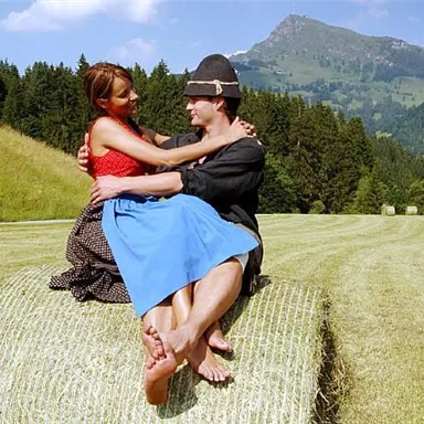 A couple is sitting on a hay bale in a green meadow with mountains in the background. They are smiling at each other and enjoying the beautiful landscape.