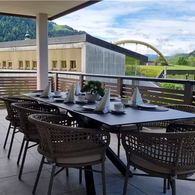 A cozy dining area on a terrace with a large table and chairs. In the background, green mountains and a blue sky can be seen.