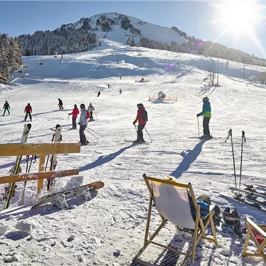 Eine schneebedeckte Skipiste mit Skifahrern und einem sonnigen Himmel. Im Vordergrund stehen Liegestühle und Skiausrüstung.