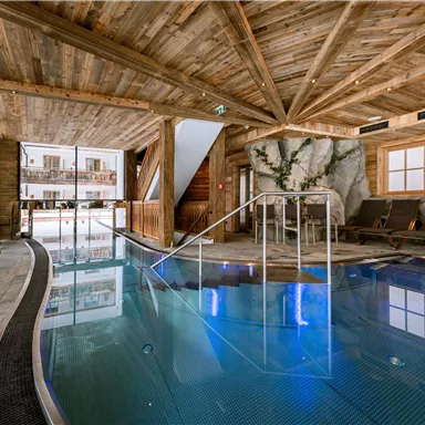 Ein eleganter Innenraum mit einem glitzernden Pool und gemütlichen Liegen. Die Wände und Decke aus Holz verleihen eine warme Atmosphäre.