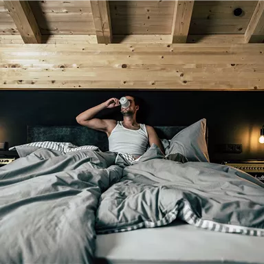 Ein Mann sitzt im Bett und trinkt aus einer Tasse. Das Zimmer hat eine moderne Einrichtung mit einem Holzofen und einer gemütlichen Atmosphäre.