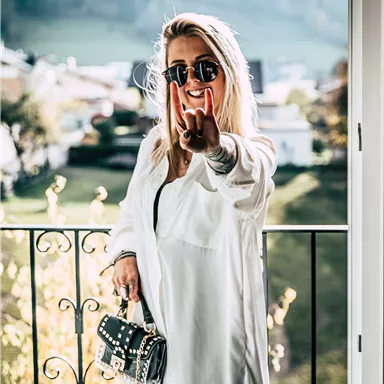 Eine lächelnde Frau mit Sonnenbrille steht auf einem Balkon und zeigt das Peace-Zeichen. Hinter ihr ist eine grüne Landschaft mit Bäumen und Hügeln zu sehen.