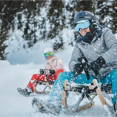 Zwei Personen fahren auf Schlitten durch den Schnee. Sie haben Winterkleidung und Helme an und genießen die verschneite Landschaft.