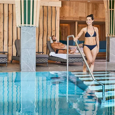 Ein moderner Wellnessbereich mit einem klaren Pool. Eine Frau in einem Bikini tritt ins Wasser, während ein Mann im Hintergrund entspannt.