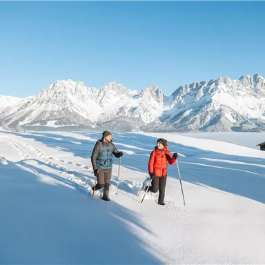 Zwei Menschen wandern mit Schneeschuhen durch eine verschneite Landschaft. Im Hintergrund sind beeindruckende Berge und ein klarer blauer Himmel zu sehen.