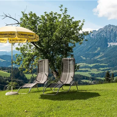 Ein ruhiger Garten mit zwei Liegestühlen und einem Gelb-weißen Sonnenschirm. Im Hintergrund sind majestätische Berge und eine grüne Landschaft zu sehen.
