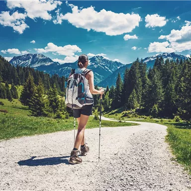 Eine Wanderin geht auf einem Kiesweg durch die Berge. Im Hintergrund sind hohe Berge und ein blauer Himmel mit weißen Wolken zu sehen.