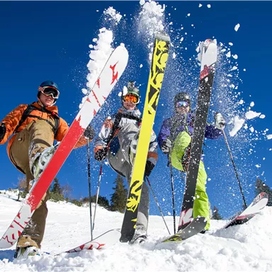 Three skiers are standing in the snow and showing their skis. The sky is clear and blue, and fresh snow is being stirred up.