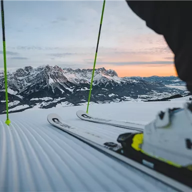 A ski view of snow-covered mountains in the background. In the foreground, skis and a ski boot can be seen.