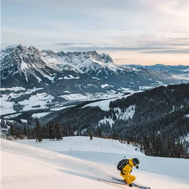 Ein Skifahrer in gelber Kleidung fährt eine Piste hinunter. Die Berge und das verschneite Tal sind im Hintergrund sichtbar.