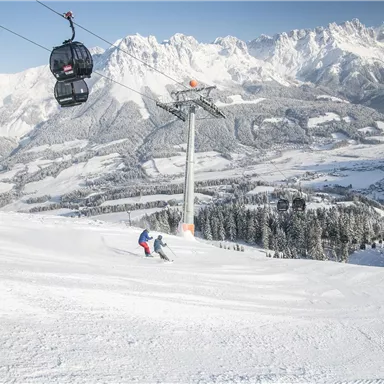 Eine verschneite Berglandschaft mit Skifahrern auf der Piste. Im Hintergrund ist eine Gondelbahn und majestätische Berge sichtbar.