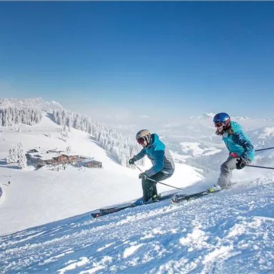 Two skiers are skiing down the snowy slope. In the background, there are snow-covered mountains and a blue sky.