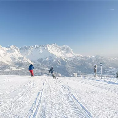 Two skiers are skiing on a snowy slope with impressive mountain peaks in the background. The sky is clear and sunny.