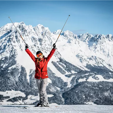 A person stands on a snow-covered mountain, lifting the ski poles into the air. In the background, impressive, snow-covered mountains can be seen.