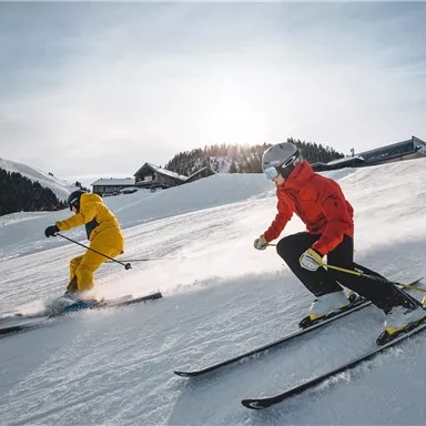 Two skiers are descending the slope, one in a yellow suit and the other in a red suit. The winter landscape is covered in snow and showcases the beauty of the mountains.