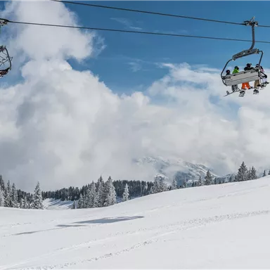 A snow-covered landscape with a ski lift and clouds in the sky. In the foreground, you can see untouched snow and trees.