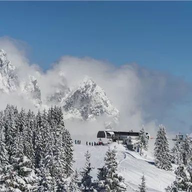 Eine malerische Schneelandschaft mit hohen Bergen und einem klaren blauen Himmel. Im Vordergrund sind schneebedeckte Bäume und eine Bergstation zu sehen.