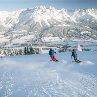 Two skiers glide over a snowy slope. In the background, majestic mountains stretch under a clear sky.