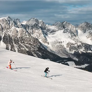Two skiers are going down the snowy slope. In the background, impressive mountains can be seen under a gray sky.