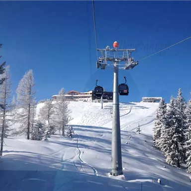 Eine Seilbahn fährt über schneebedeckte Berge. Im Hintergrund sind verschneite Bäume und ein Gebäude sichtbar.