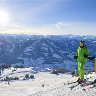 Zwei Skifahrer stehen auf einem Berg mit schneebedeckten Hängen. Im Hintergrund sind majestätische Berge und ein klarer Himmel zu sehen.