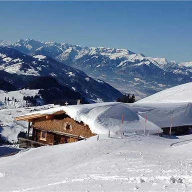 Eine verschneite Berglandschaft mit einem traditionellen Holzhaus. Im Hintergrund sind majestätische Berge und ein klarer blauer Himmel zu sehen.