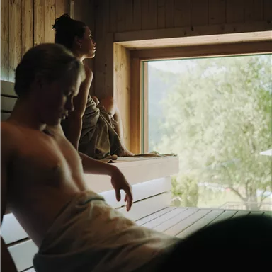 Zwei Personen entspannen in einer Sauna. Der Raum ist aus Holz und bietet einen Blick auf die Natur draußen.