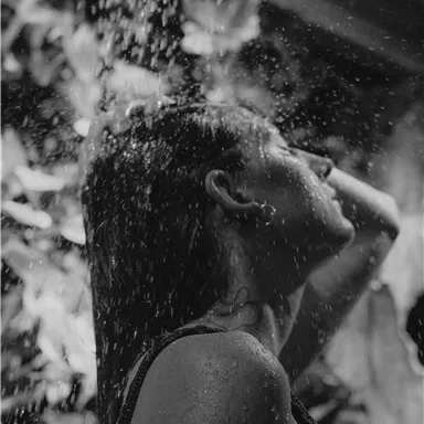 Eine Frau steht unter der Dusche und genießt das Wasser. Die Szene ist in Schwarz-Weiß gehalten, mit einer natürlichen Umgebung im Hintergrund.