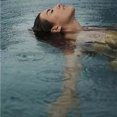 Eine Person liegt entspannt im Wasser, während es regnet. Ihr Gesicht zeigt Zufriedenheit und Ruhe.