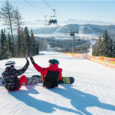 Zwei Snowboarder sitzen im Schnee und feiern gemeinsam. Im Hintergrund sind schneebedeckte Berge und eine Seilbahn zu sehen.