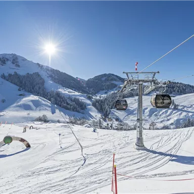 An idyllic winter landscape with skiing slopes and a chairlift. The sky is clear and the sun is shining over the snow-covered mountains.