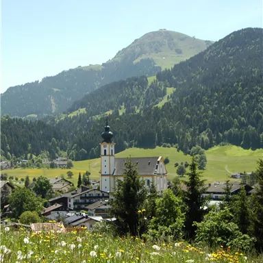 Eine malerische Landschaft mit einem Dorf und einer Kirche im Vordergrund. Im Hintergrund sind grüne Berge und ein klarer blauer Himmel zu sehen.