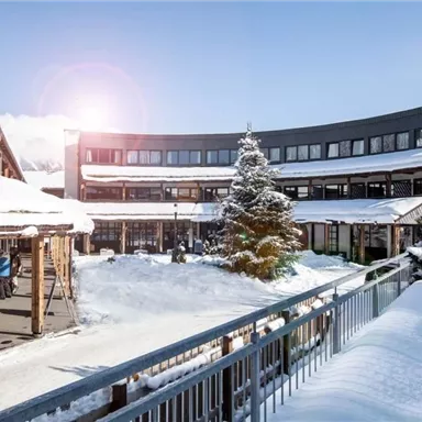 Ein malerischer Innenhof mit schneebedecktem Boden und einem Weihnachtsbaum. Die moderne Architektur umgibt den Raum, während die Sonne scheint.