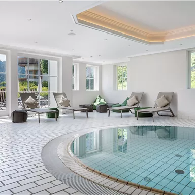A bright wellness room with a round pool and lounges. Large windows offer a view of the surrounding nature.