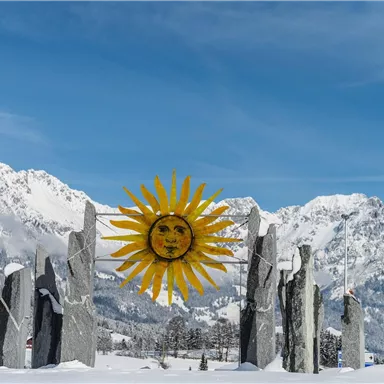 A radiant sun chariot made of stone in a snowy landscape. In the background, majestic mountains are visible under a blue sky.