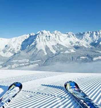 A snow-covered mountain landscape with a clear blue sky. In the foreground, two skis are lying on the groomed slope.