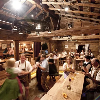 Eine gemütliche Almwirtschaft mit Holzinterieur. Menschen in traditioneller Kleidung tanzen und feiern.