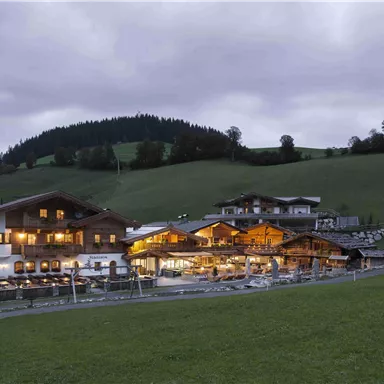 Ein typisches alpenländisches Gebäude in der Dämmerung, umgeben von grünen Wiesen und sanften Hügeln. Die Fenster strahlen warmes Licht aus und schaffen eine einladende Atmosphäre.