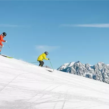 Two skiers are skiing down the snowy slope. In the background, impressive mountains and a blue sky can be seen.