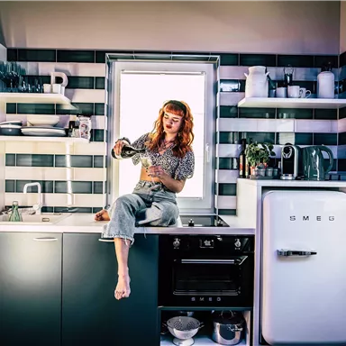 Eine moderne Küche mit einer Frau, die auf der Arbeitsplatte sitzt und Wasser aus einem Krug gießt. Die Wände sind in stilvollen Farben gestaltet und die Fenster bieten viel Licht.