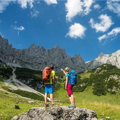 A pair of hikers stands on a rock, looking at majestic mountains. The sky is clear and blue, with some beautiful clouds.