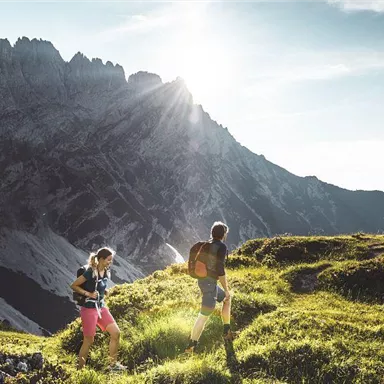 Two hikers explore a green meadow in the mountains. In the background, tall rocks are visible, and the sun is shining brightly.