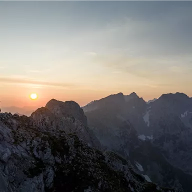 Zwei Personen stehen auf einem Berggipfel und genießen den Sonnenuntergang. Die umliegenden Berge sind in sanften Farben beleuchtet.