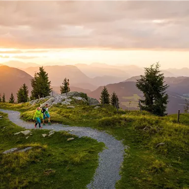 Eine malerische Berglandschaft während des Sonnenuntergangs. Wanderer folgen einem schmalen Pfad zwischen Bäumen und Bergen.